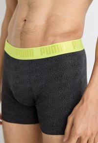 Black boxer briefs with a repeating PUMA logo pattern, featuring a bright yellow elastic waistband. Made from a smooth, stretchy fabric.