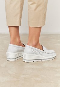Scarpe bianche slip-on con design perforato, suola a zeppa rialzata e accenti testurizzati. Sfondo neutro con pantaloni beige a vita alta.
