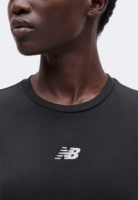 Κοντινό πλάνο ενός ατόμου που φοράει μαύρο T-shirt New Balance, δείχνοντας τον λαιμό, το κάτω μέρος του προσώπου και το λογότυπο στο στήθος.