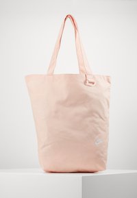 Jasnoróżowa torba na ramię Nike Air z długimi uchwytami, bia Logo nadrukowane z przodu i duży pionowy tekst "NIKE" z boku.