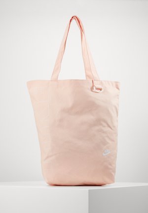 Sac fourre-tout Nike Air rose clair avec de longues poignées, logo blanc imprimé à l'avant et grand texte vertical "NIKE" sur le côté.