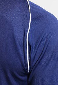 Marinblå träningsskjorta med ett strukturerat stickat mönster, med vita detaljer längs axelsömmen. Andningsbart material.