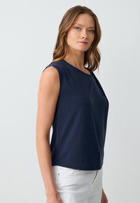 Top senza maniche di colore blu navy in tessuto morbido; presenta una scollatura rotonda e un dettaglio di drappeggio sottile su un lato; abbinato a pantaloni bianchi.