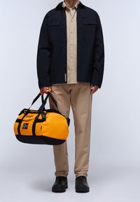 Giacca nera con due tasche sul petto, camicia beige, pantaloni color camoscio e scarpe nere. Tiene in mano una borsa da viaggio arancione e nera con un logo e una bandiera.