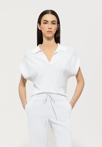 Weißes Strickoberteil mit V-Ausschnitt und kurzen Fledermausärmeln, kombiniert mit hellen, lässigen Hosen mit Kordelzug. Glatte Textur, minimalistisches Design.