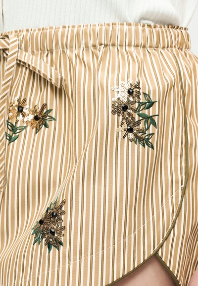 Μπεζ και λευκή ριγέ φούστα τύπου wrap με λεπτομέρειες από φλοράλ παγιέτες και κεντημένα πράσινα φύλλα σε άτομο που φοράει λευκή μπλούζα.