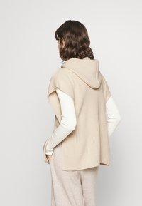 Poncho lavorato a maglia beige con cappuccio. Presenta maniche corte e una vestibilità ampia. Indossato sopra un top bianco a maniche lunghe, con una texture morbida.