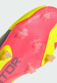 Semelle de chaussure de football en rose et jaune, présentant une surface texturée et des crampons translucides conçus pour offrir une traction sur les surfaces en herbe.