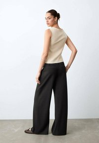 Femeie cu părul prins într-un coc, purtând un top fără mâneci bej, pantaloni negri cu croială largă și pantofi maro fără toc, stând în picioare pe un fundal simplu, deschis la culoare.