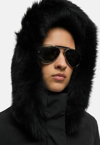 Giacca parka nera con cappuccio foderato in pelliccia spessa. Presenta occhiali da aviatore riflettenti e una texture di tessuto liscia.