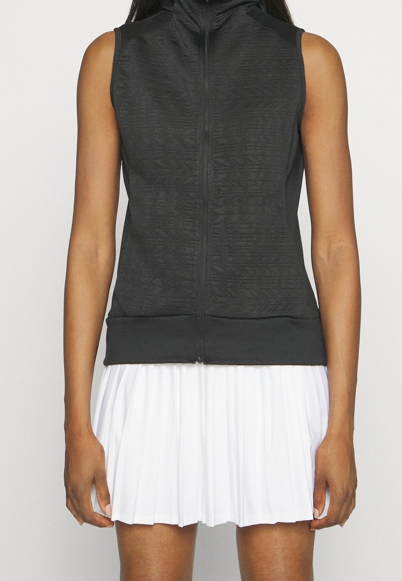 Sudadera sin mangas negra con cremallera, con un patrón texturizado, cuello alto y un diseño ajustado. Combinada con una falda blanca con plisados.