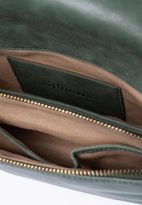 Groene leren handtas interieur met lichtbruine voering, rits met gouden hardware en een label dat aan de binnenkant is gestikt. Eenvoudig, functioneel design.