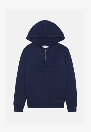 Tumši zila kapucēm jaka ar rāvējslēdzēju pie kakla. Iespējas ar ribotām aprocēm un apakšmalu, izgatavota no mīkstas adīta materiāla. Ādas zīmola etikete pie apakšmalas.