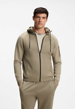 Mand iført khaki lynlåshoodie med sorte snore og matchende bukser, stående med hænderne i lommerne mod hvid baggrund.