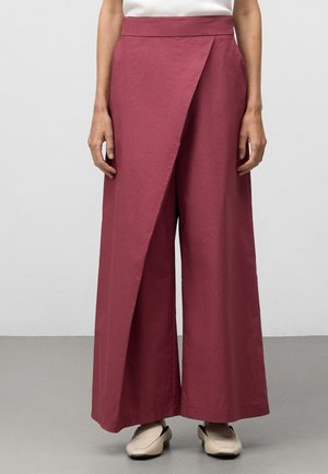 Nainen pukeutunut leveälahkeisiin, korkeavyötäröisiin liilan sävyisiin housuihin, joissa on päällekkäinen etupaneeli, ja beigen värisiin neliskanttavartisiin kenkiin, seisoo harmaalla lattialla.