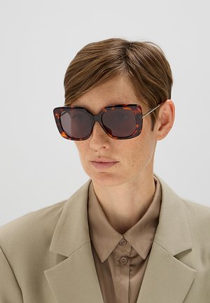 Persona con capelli castani corti che indossa grandi occhiali da sole quadrati con montatura tartarugata e un blazer beige sopra una camicia abbottonata.