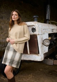 Beżowy sweter wełniany w połączeniu z jasnobeżową spódnicą w kratę z zielonymi akcentami. Stojąc obok rustykalnego, białego traktora z metalowymi detalami.