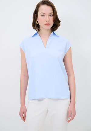 Lyseblå, kortærmet bluse med V-hals og krave. Glat tekstur, minimalistisk design. Bæret med hvide bukser.
