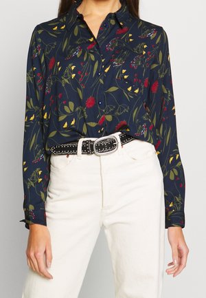 Femme porte un chemisier bleu marine à manches longues avec un motif floral et d'oiseaux multicolore, rentré dans un pantalon blanc taille haute et une ceinture noire cloutée.