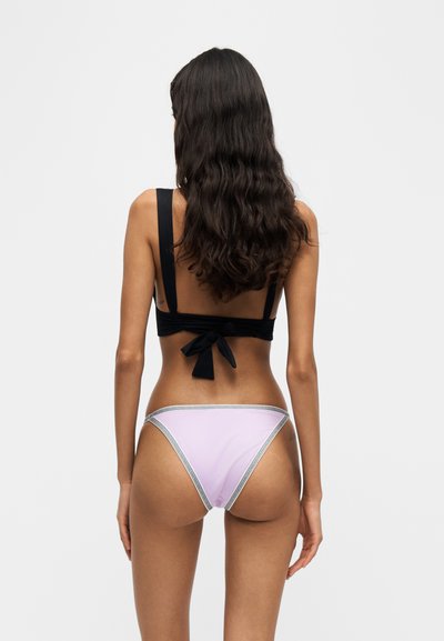 Femme aux longs cheveux foncés porte un haut de bikini noir noué dans le dos et un bas de bikini violet clair avec un bord contrasté, vue de dos.