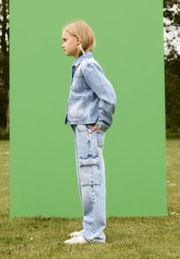 Jeansjacka och cargobyxor i ljusblå tvätt, med stora fickor och en avslappnad passform, kompletterade av vita skor och en grön bakgrund.