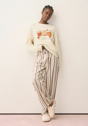 Kvinde iført cremefarvet sweatshirt med illustration af efterårsdrikke, løse stribede bukser og flagrende tøfler, stående op af en ensfarvet væg.