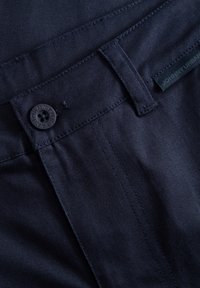 Námořnické modré džíny s hladkou texturou, které mají kulatý černý knoflík, poutka na opasek a malou značkovou nálepku blízko pasu.