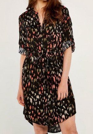 Kobieta w czarnej sukience z wielokolorowymi abstrakcyjnymi wzorami, z podwiniętymi rękawami, zapinanej na guziki z przodu, z przewiązanym paskiem w talii.