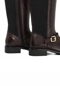 Brown leather riding boots with a black elastic panel, side zipper, and gold buckle accent on the strap above the heel.