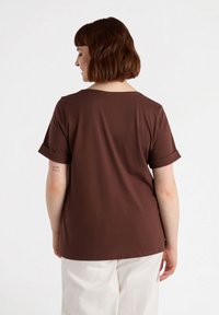 Camiseta marrón de manga corta con un corte relajado, un cuello redondo amplio y puños enrollados, combinada con pantalones de color claro, vista desde la parte de atrás.