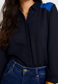 Camiseta azul marina con cuello abotonado, que presenta paneles de acento azul y naranja en los hombros. Llevada con jeans de mezclilla azul oscuro.