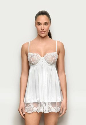 Woman with dark hair in a ponytail wearing white lace and satin lingerie dress, standing against a plain light background, facing forward.