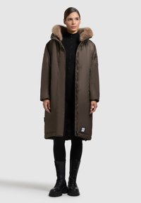 Kvinna iförd en lång brun vadderad kappa med foder av päls, svarta leggings och svarta snörade stövlar, stående mot en enfärgad bakgrund.