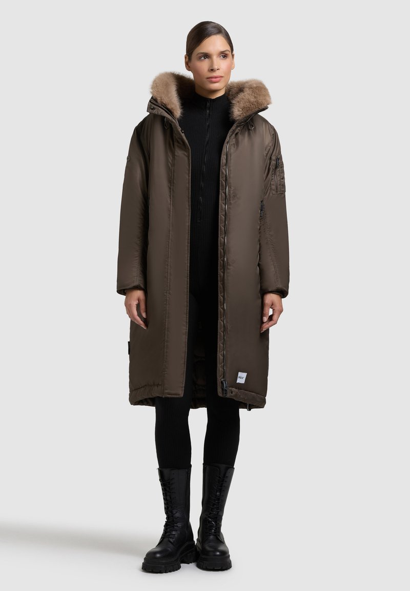 Kvinna iförd en lång brun vadderad kappa med foder av päls, svarta leggings och svarta snörade stövlar, stående mot en enfärgad bakgrund.