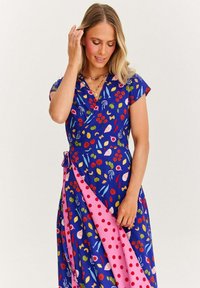 Modré zavinovací šaty s květinovým vzorem v červené, žluté a zelené; růžová sukně s bílými puntíky. Krátké rukávy.