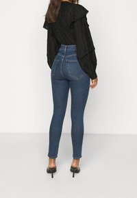 Jeans de mezclilla azul de tiro alto con una silueta ajustada, costuras detalladas y dos bolsillos traseros; combinados con una blusa negra texturizada.