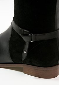 Bottine en daim noir et cuir avec une sangle décorative en cuir et un rivet argenté, dotée d'un petit talon bas en bois empilé.