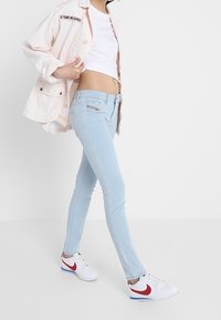 Jeans ajustados de color azul claro con un ajuste de tiro medio, combinados con una camiseta corta blanca y una chaqueta rosa. Zapatillas blancas con detalles rojos completan el look.