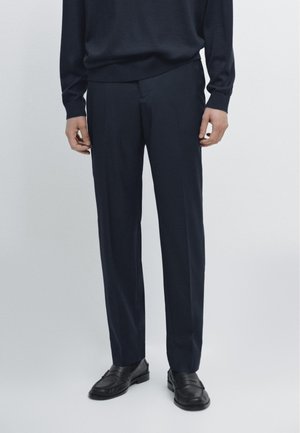 Man wearing black dress pants, black long-sleeve shirt, and black loafers standing against a plain light background.