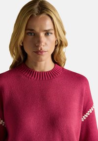 Pulover tricotat roz cu un guler cu nervuri, dotat cu accente decorative albe pe mâneci, oferind o textură moale și o croială lejeră.