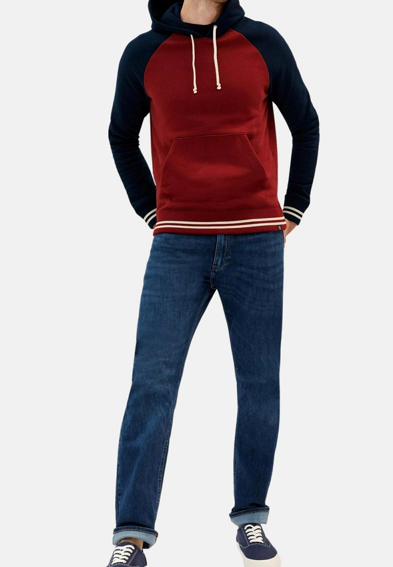 Homme portant un sweat à capuche rouge et bleu marine avec des cordons blancs, un jean bleu retroussé et des baskets bleu marine à semelles blanches.