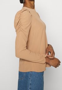 Camicia beige a maniche lunghe con dettagli arricciati sulle spalle e sulle maniche, realizzata in un tessuto morbido, abbinata a jeans blu a vita alta.