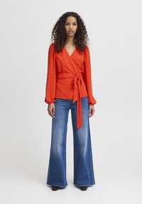 Blouse rouge à manches longues avec détail à nouer à la taille, associée à un jean en denim bleu évasé. Tissu lisse avec un motif texturé.