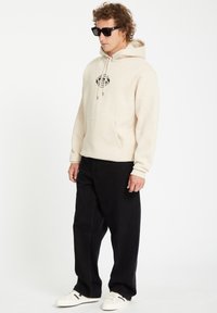Beige hoodie met een zwart grafisch logo op de borst, gecombineerd met zwarte jeans. Witte sneakers met zwarte accenten maken de outfit compleet.