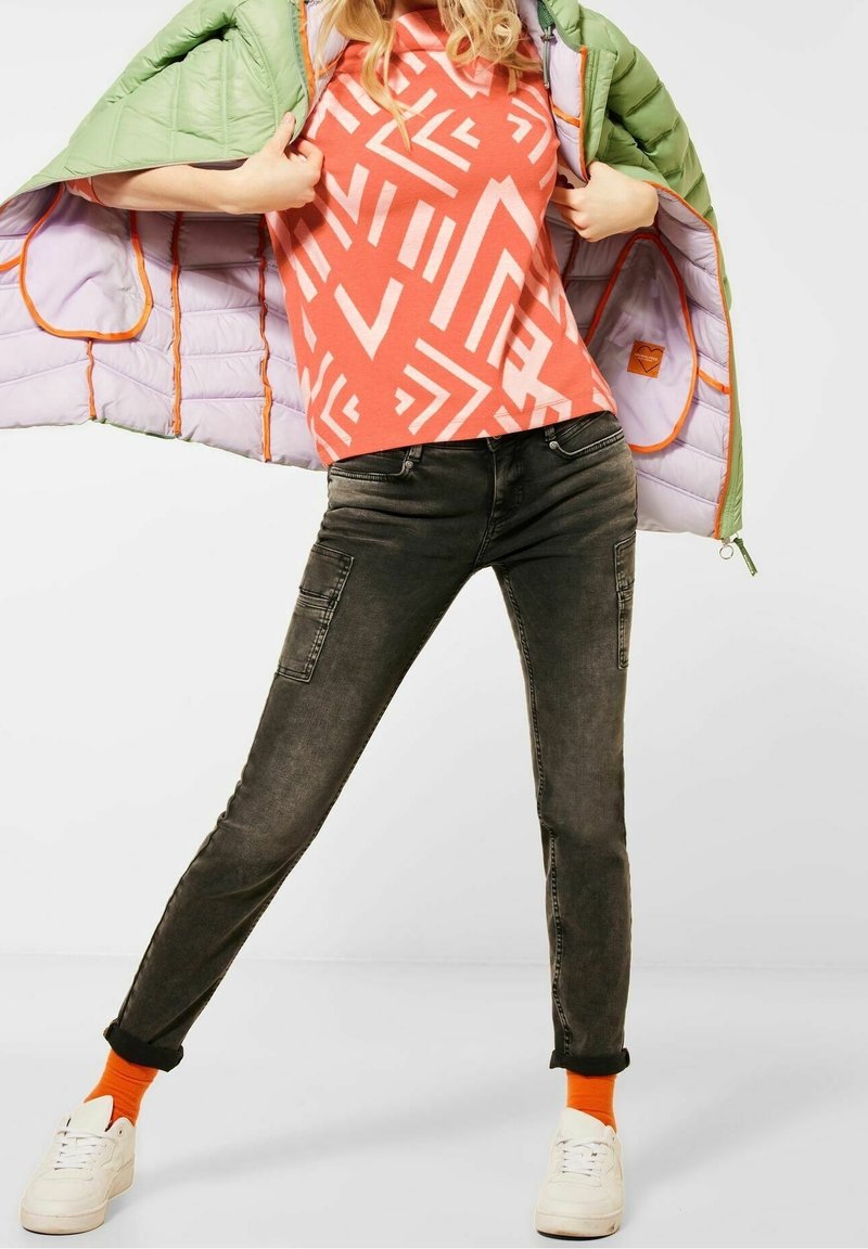 Green puffer jacket over a coral patterned shirt, dark denim cargo pants, white sneakers, and orange socks. Smooth textures, modern fit.