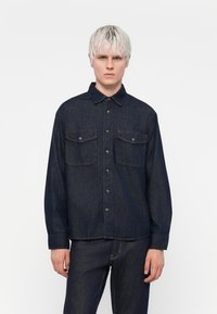 Mørk denimskjorte med to framlommer, knappelukking, spiss krage og lange ermer. Glatt tekstur med kontraststing.
