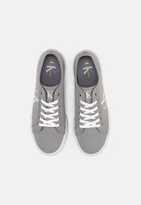 Graue Leinenschuhe mit weißen Schnürsenkeln, runder Zehenpartie, Gummisohle und einem grünen Logo auf den Innensohlen. Schlichtes Design mit minimalen Details.