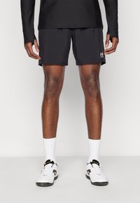 Μαύρο αθλητικό σορτς από αεριζόμενο ύφασμα, εφαρμοστό σχέδιο, μήκος πάνω από το γόνατο, συνδυασμένο με λευκές κάλτσες και μαύρα αθλητικά παπούτσια. Ορατό το λογότυπο της Under Armour.