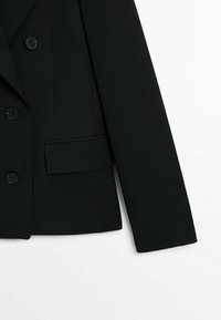 Blazer noir à manches longues, trois boutons et une poche à rabat sur un fond blanc uni.