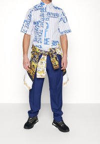 Kortärmad skjorta med knappar och blå logotryck, kombinerad med marinblå byxor. En mönstrad sidenhalsduk är knuten runt midjan. Svarta sneakers.